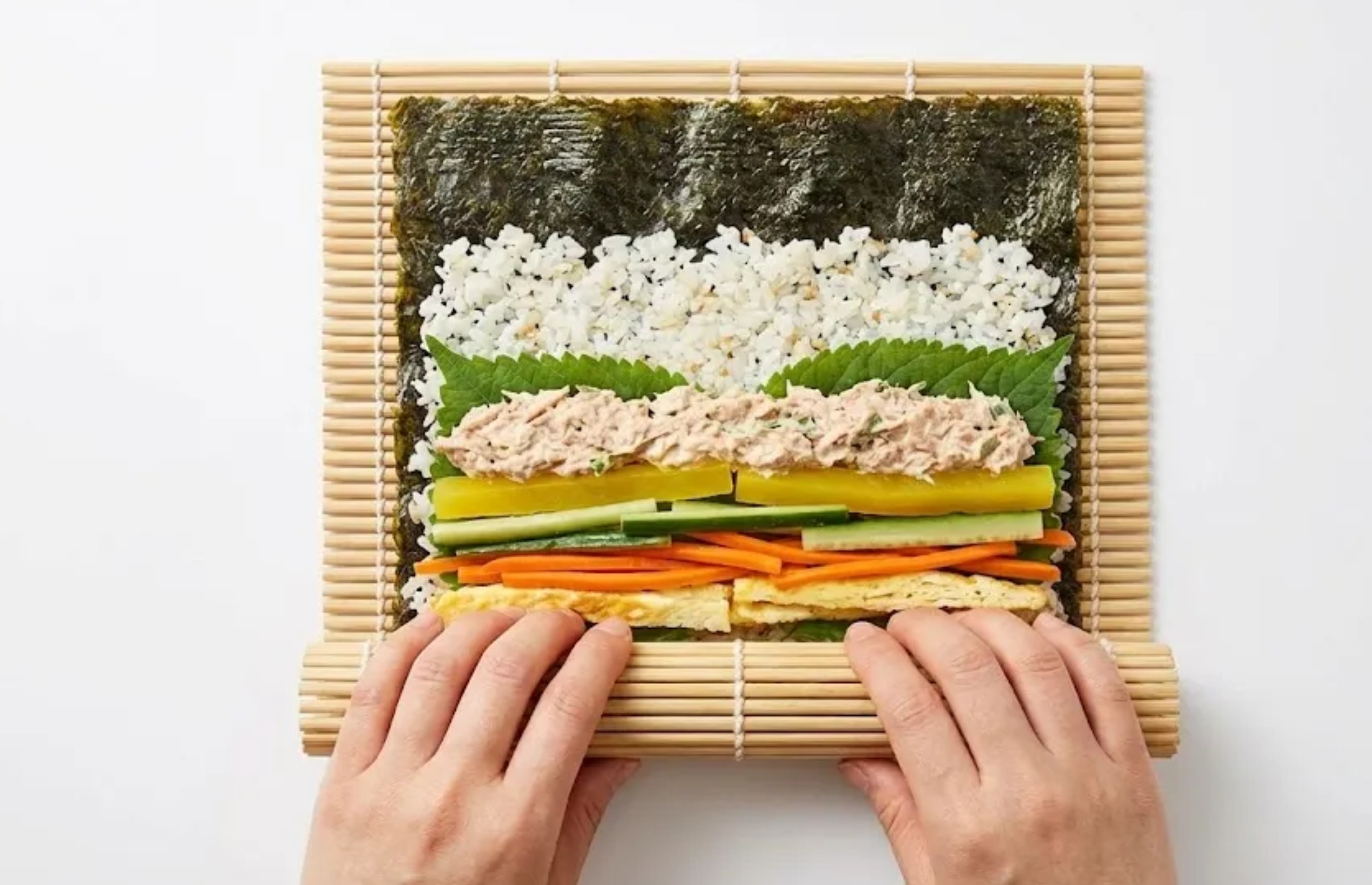 Hands rolling kimbap on a bamboo mat with colorful fillings of carrot, spinach, egg and ham inside
